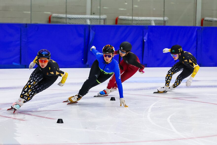 A short-track speed skating race with multiple athletes leaning into a turn on the ice.