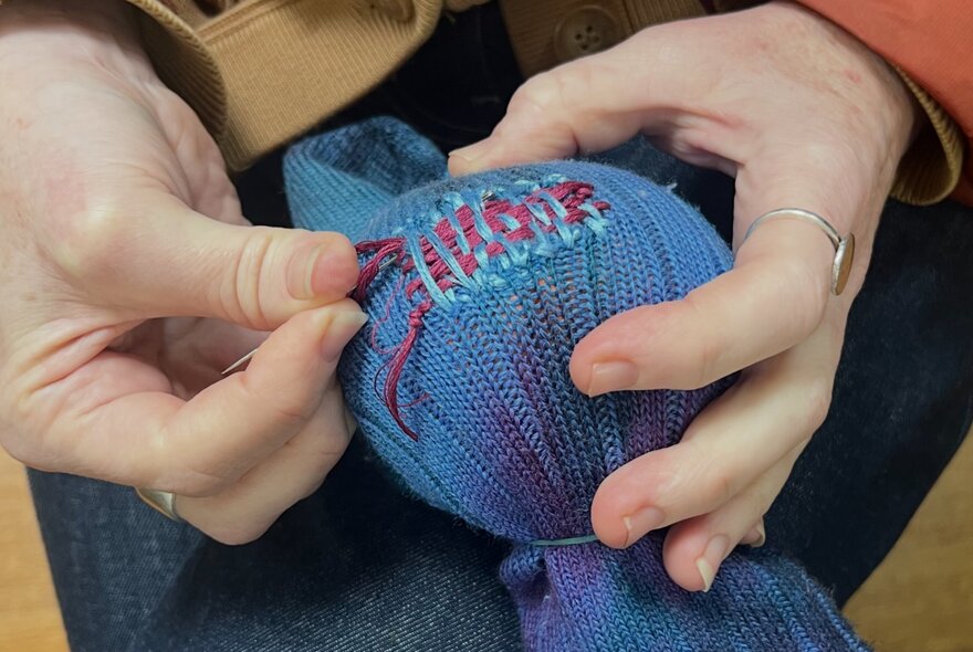 A pair of hands using a darning needle with red thread, to repair the sleeve of a blue knitted jumper. 