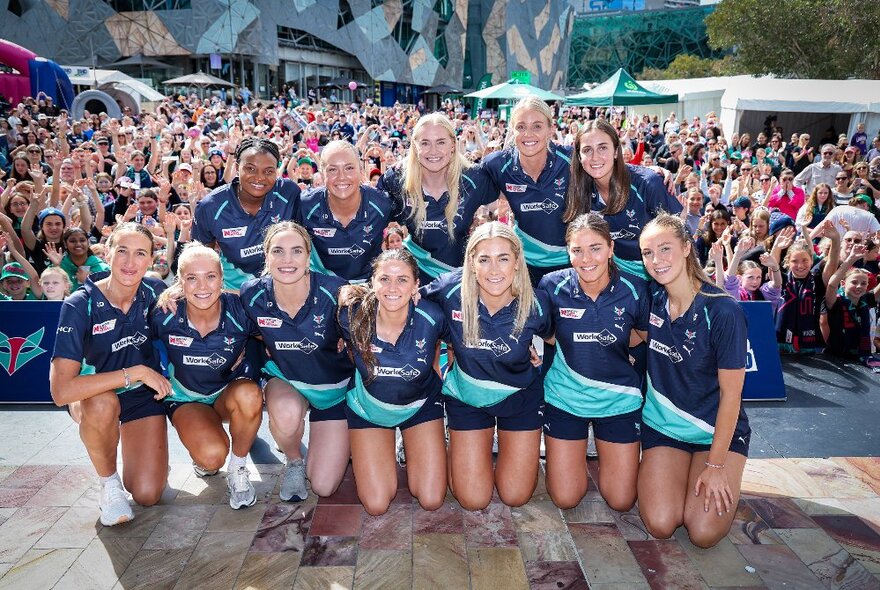 Vixens netball team posing in two rows of players, the bottom row kneeling on the ground, in front of packed crowds in Fed Square.