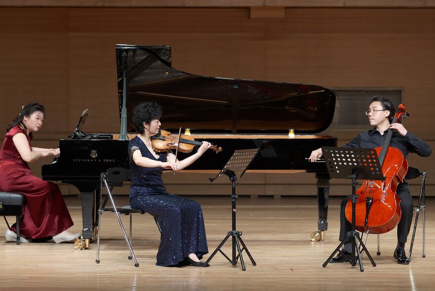 Three musicians, one playing a grand piano, another violin and the third, cello, on stage.