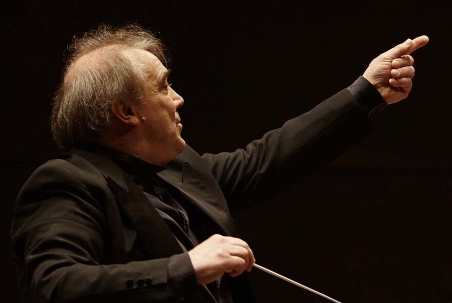 Classical conductor wearing black tux, holding a baton and pointing with one finger.