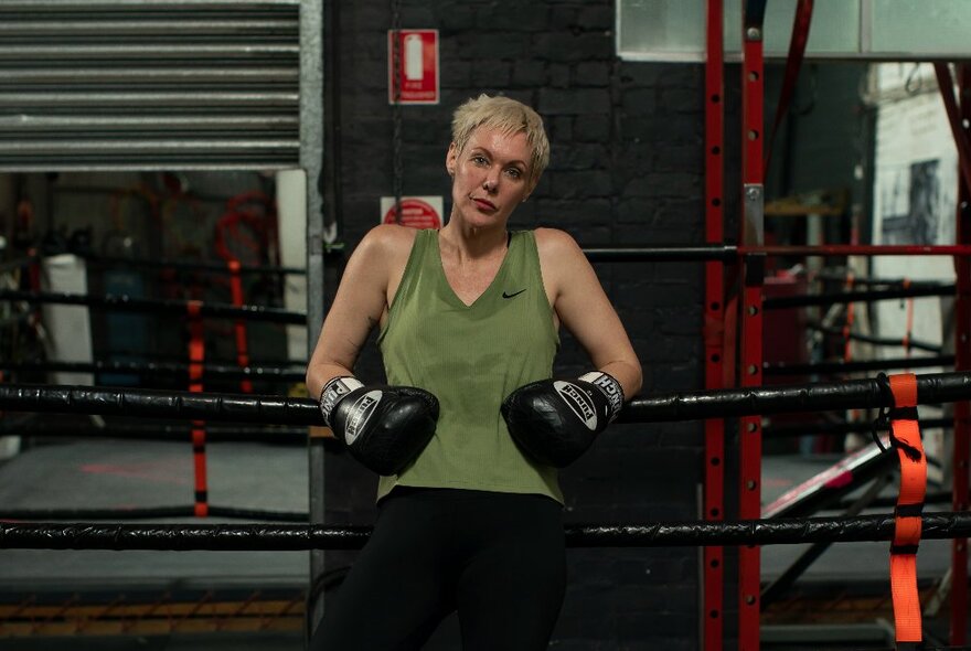 A woman wearing boxing gloves and a green singlet leaning back on the ropes of a boxing ring inside a boxing gym.