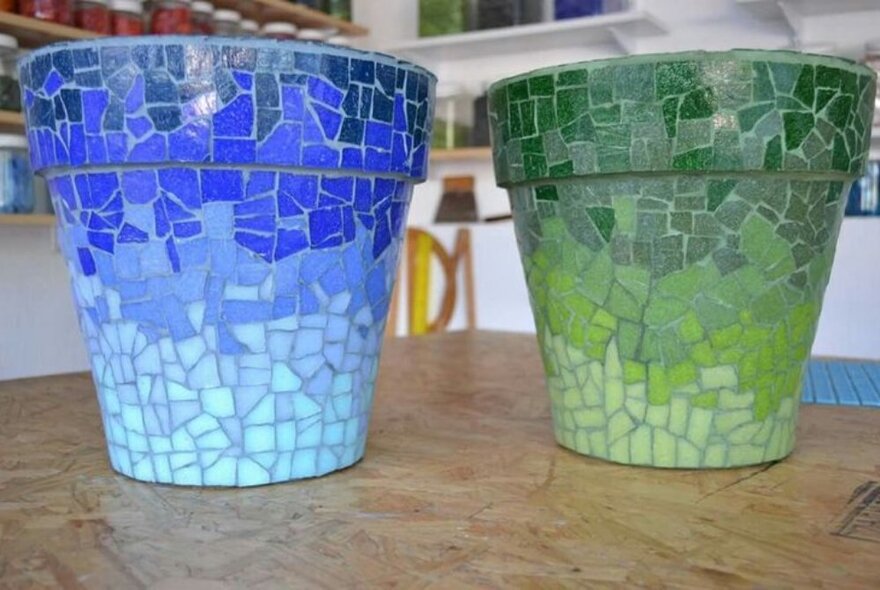 A blue and a green mosaic-tiled planter pot standing on a table in a shop.