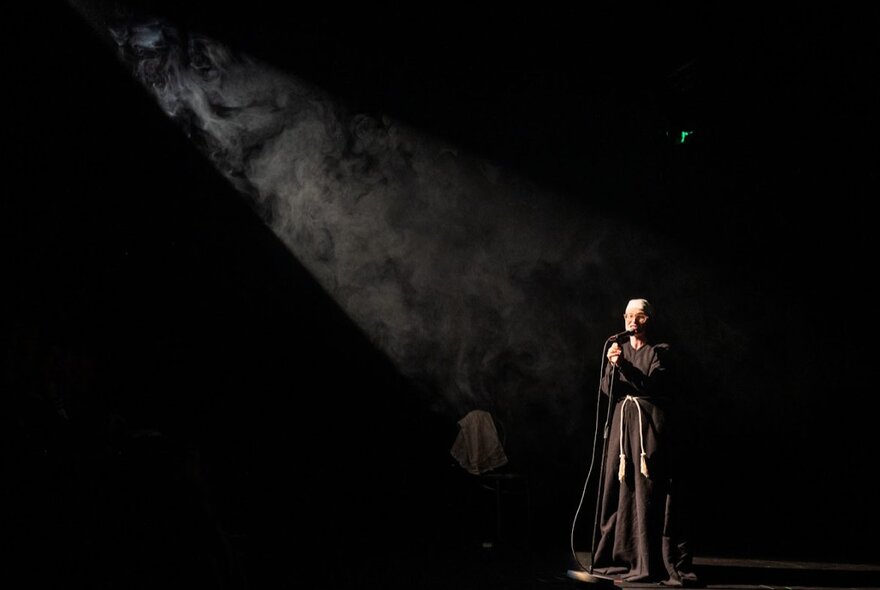 Comedian Scout Boxall on a darkened stage wearing a nun's habit and talking into a microphone.