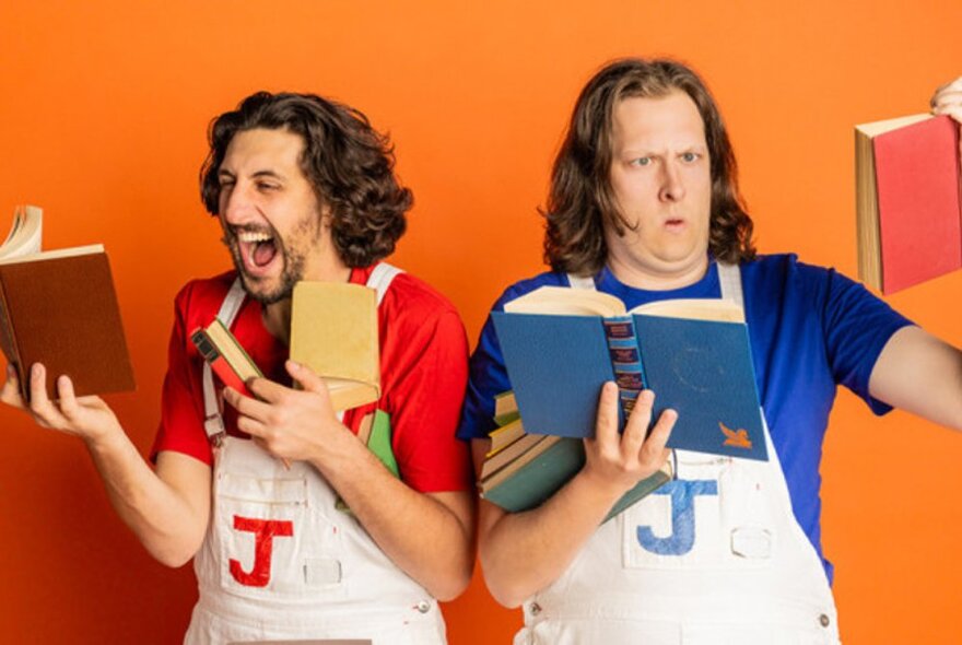 Two comedians in white overalls with bemused and silly expressions on their faces, holding multiple books in their hands and arms, standing against an orange background.