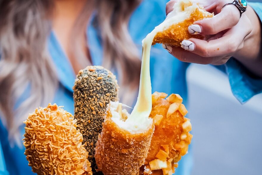 Crumbed and deep fried hot dogs on a stick, one oozing with melted cheese.