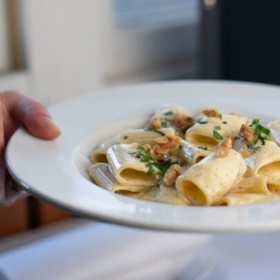 A hand holding a plate of large tubed pasta with a cream sauce and herb garnish. 