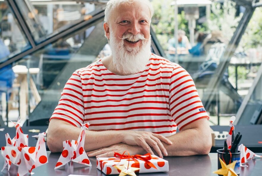 Person smiling with white hair and a white beard, wearing a red and white striped t-shirt, sitting behind hand wrapped gifts at the Atrium in Fed Square.