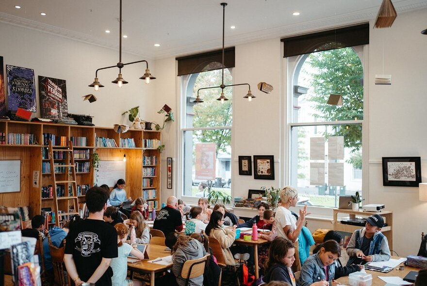 A bright and airy studio room used as a classroom, with children sitting and working around several large tables, some with laptops open in front of them, bookshelves and windows in the background.