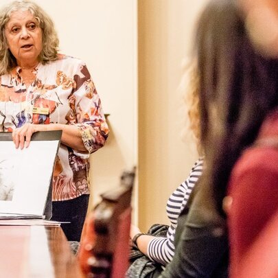 A woman presenting to a group of seated listeners, holding up a picture and making a point. 