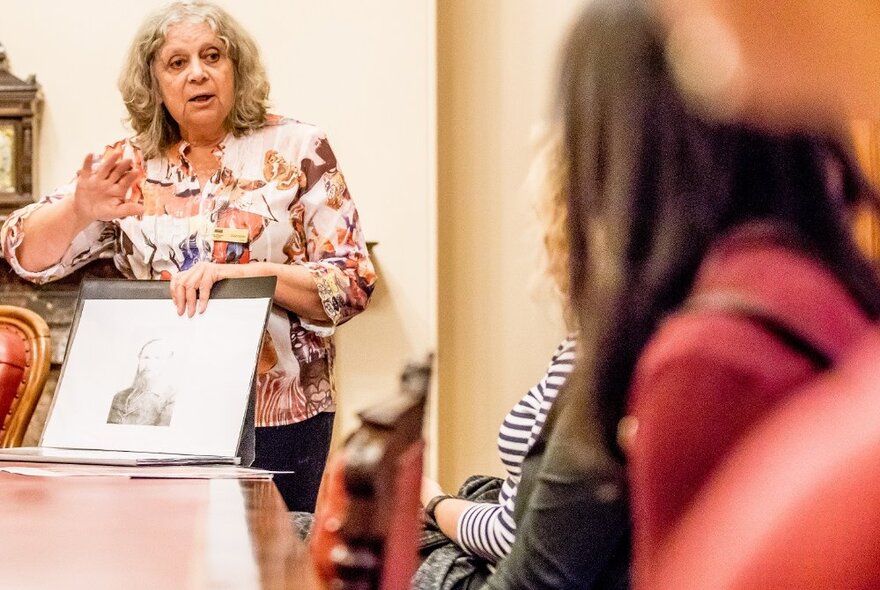 A woman presenting to a group of seated listeners, holding up a picture and making a point. 