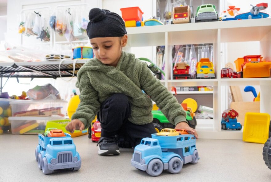 A young child seated on the floor, playing with plastic toy trucks, inside a toy library, with shelves of toys behind him.