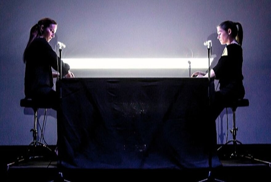 Two people seated on opposite ends of a desk, a white horizontal light between them.