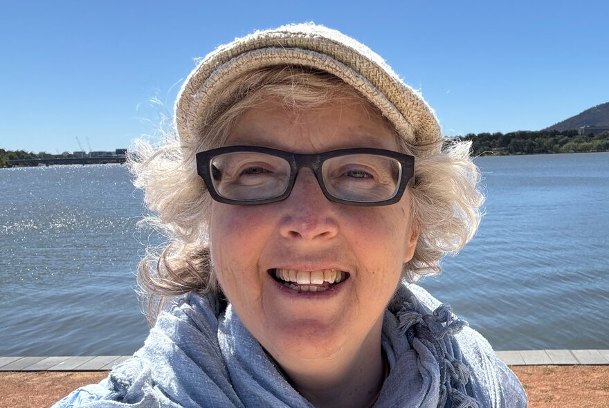 Self-portrait photograph of poet, Sarah Rice, standing in front of the ocean while wearing a hat and prescription glasses.