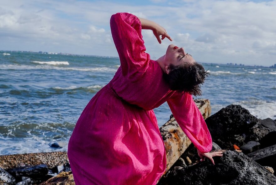 A woman in a vibrant pink dress, bending backwards at a rocky beach, the ocean waves and cloud-filled sky in the background.