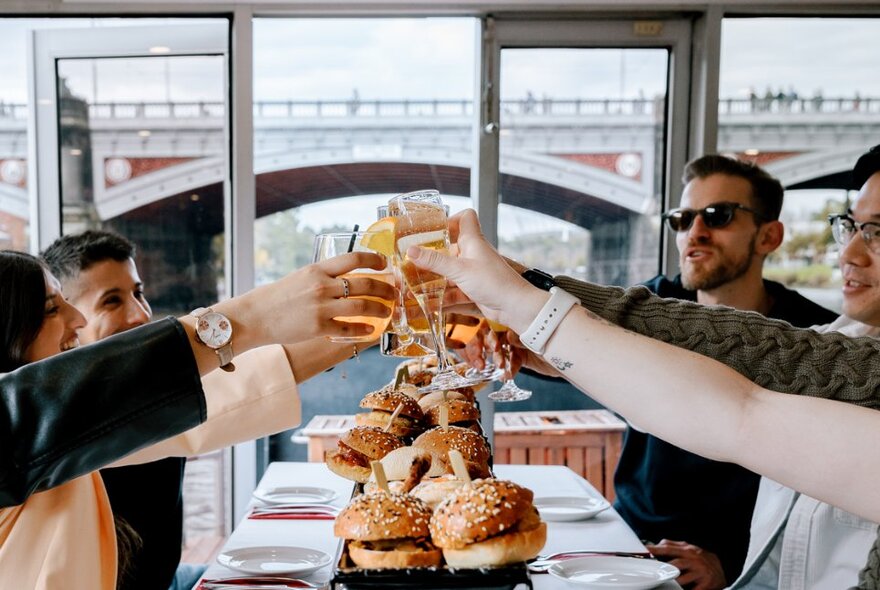 People on a long table all clinking glasses in a toast on a boat. 