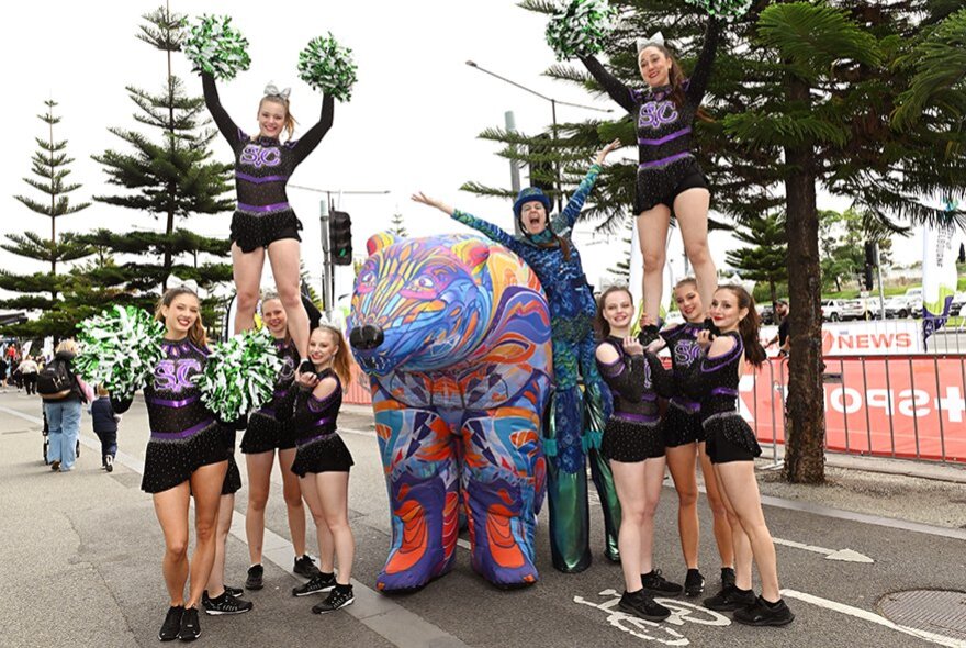 Two women standing on two groups of cheerleaders with pompoms next to a coloured animal sculpture in the city. 