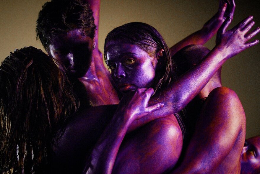 A dark image of four dancers intertwined, their bodies shining in a purple glow. 