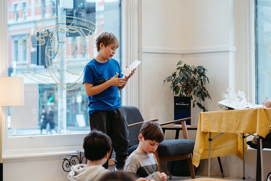 A child reading from a sheet of paper while other children sit at a table.