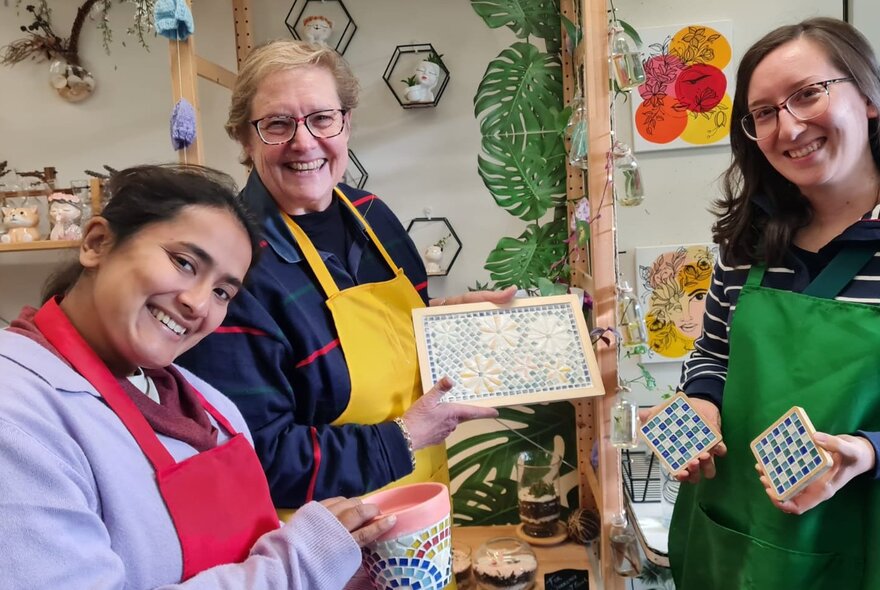 Workshop class participants smiling in a studio setting, holding up mosaic pots and coasters they have made.