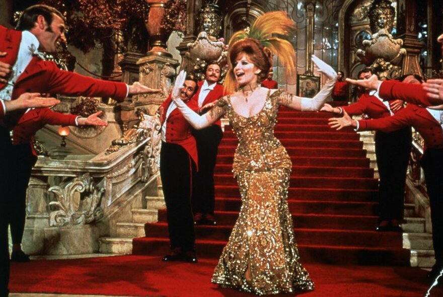 A still from the movie, Hello, Dolly! showing Barbara Streisand in a glamorous gold dress in front of red carpeted stairs; with red-jacketed bus boys reaching out to her.