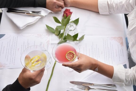 Overhead view of two people holding hands across a table with a drink in their other hand and printed menus in front of them.