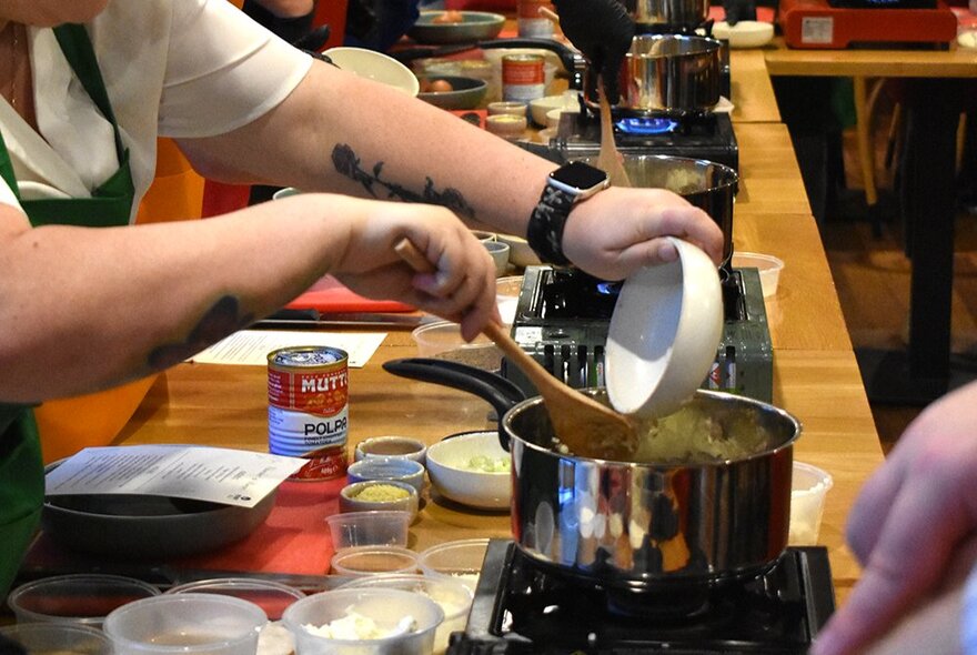 People working with pots and ingredients at a long worktable during a cooking workshop.