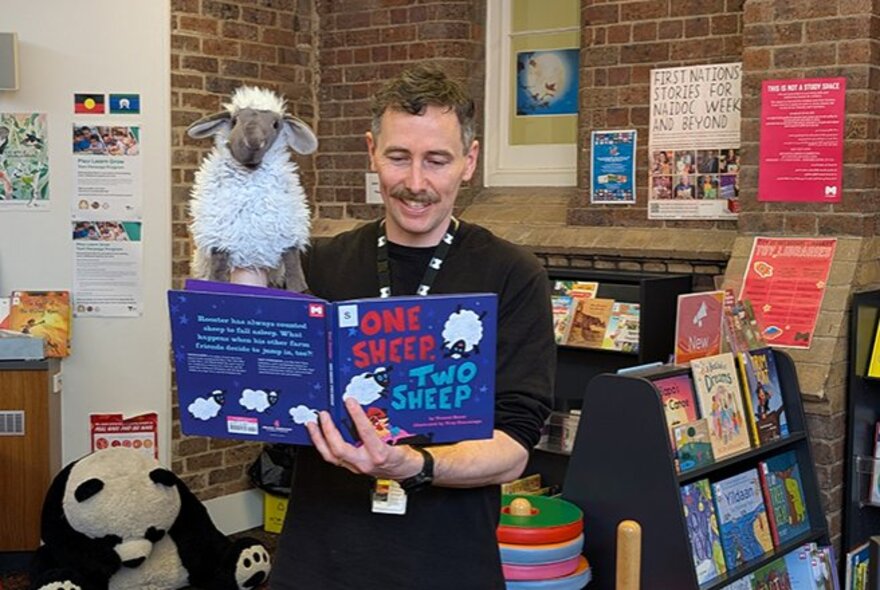 A library staff member standing in the children's book section of a library, holding up a picture book and reading from it.