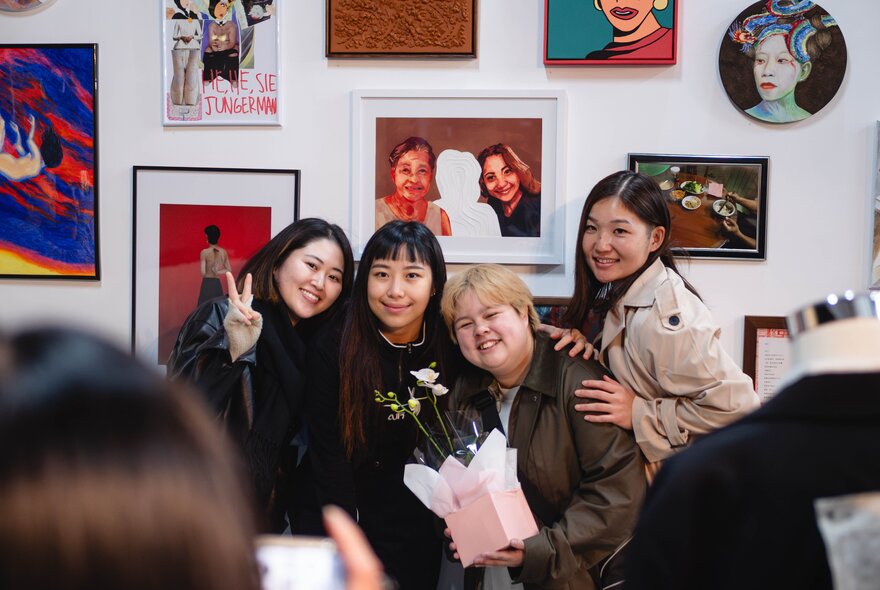 A group of friends smiling and posing for a photo in front of a wall of small artworks in a gallery space.