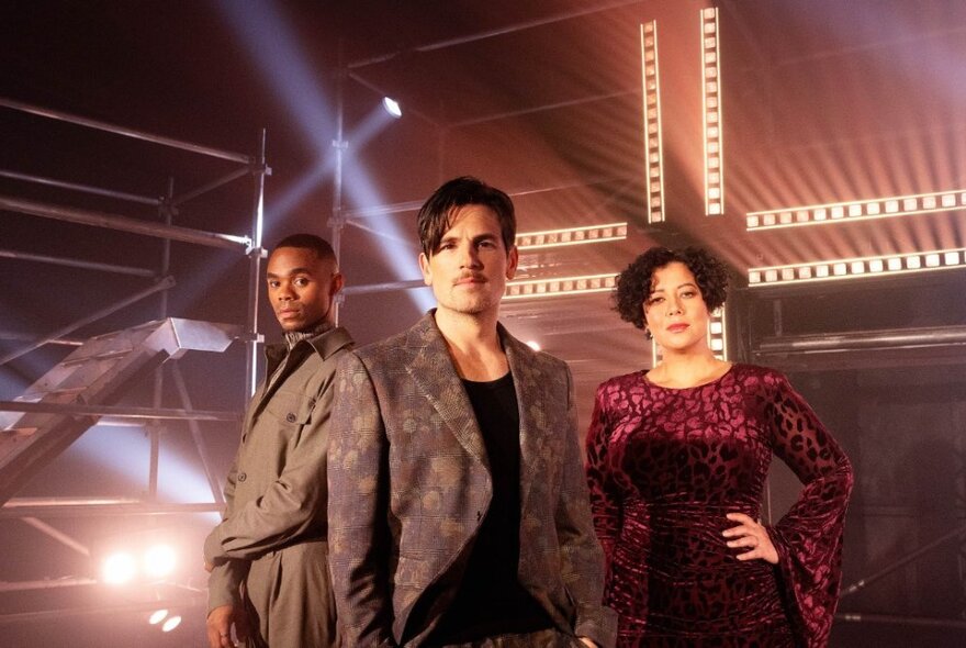 The three main cast members of the musical Jesus Christ Superstar wearing contemporary outfits in standing poses against a theatre stage backdrop.