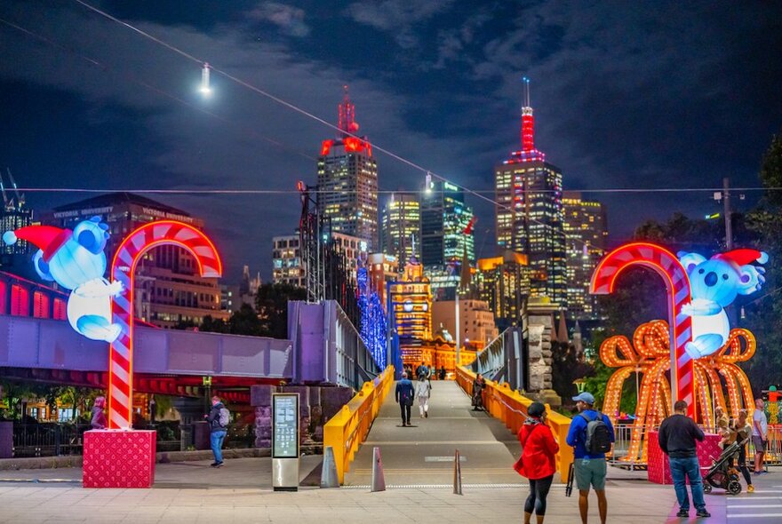 People standing around giant Christmas decorations with koalas climbing lit-up candy canes at Queensbridge Square at night, city buildings in the background.