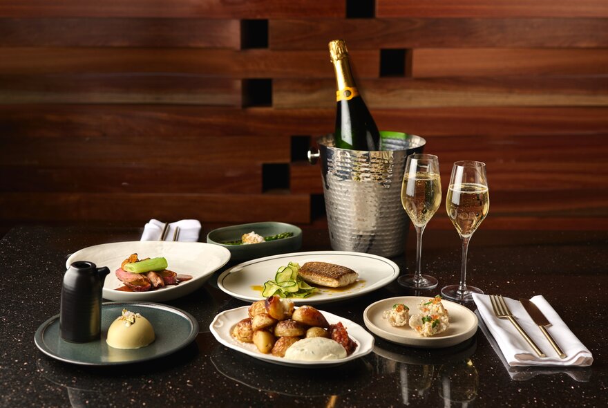 A table set wth multiple plates of carefully arranged food, a bottle of sparkling wine in and ice bucket and two glasses of bubbles. 
