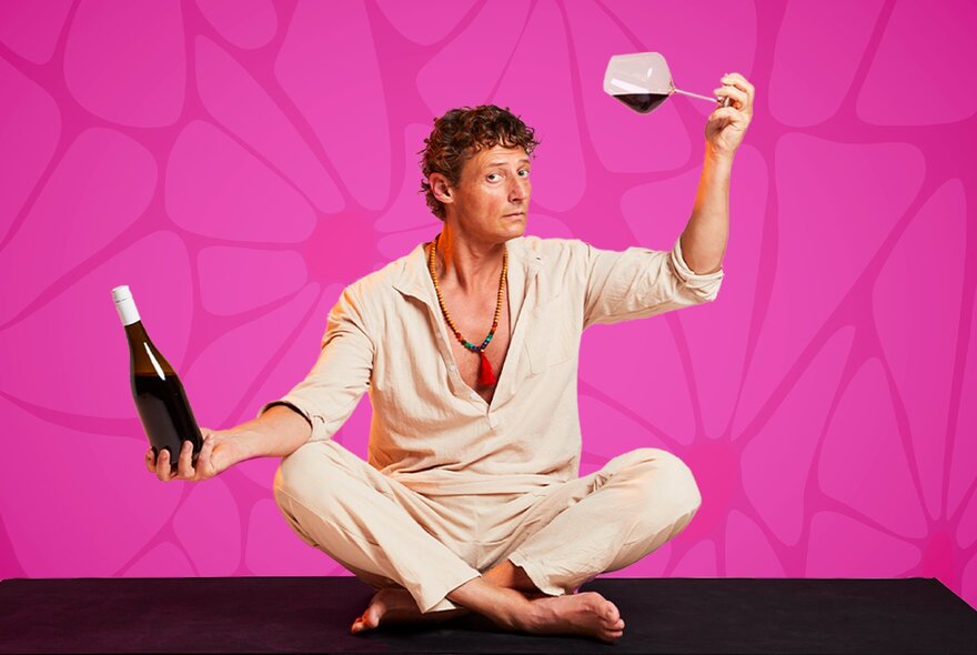 A man in linen pyjamas sitting cross legged on a black surface while holding a wine bottle in one hand and an almost horizontal wine glass in the other; pink background.