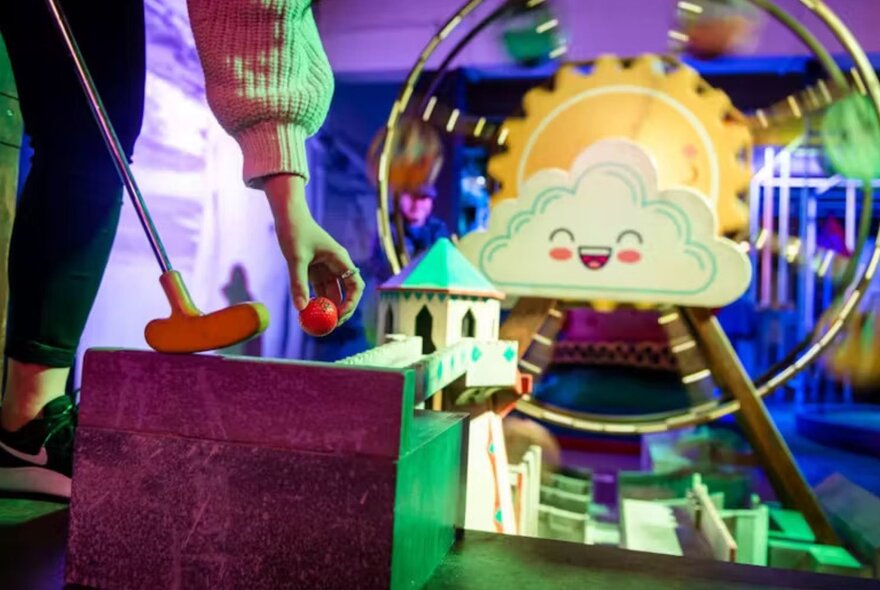 Hand holding minigolf club and ball next to cartoon-themed minigolf course.