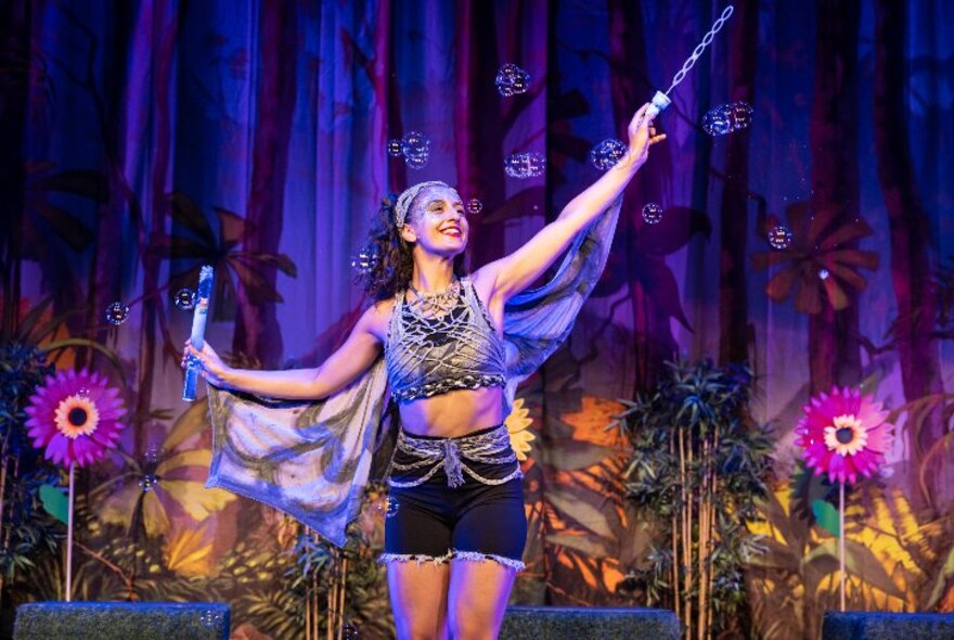 A performer on stage acting as Tinkerbell and holding up a wand with large floral stage props behind her. 