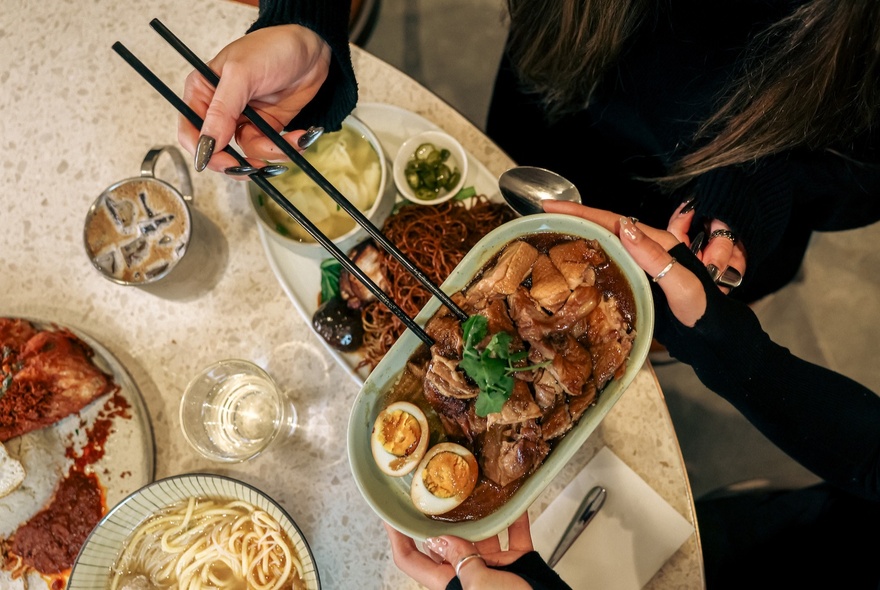 Looking down at a table with assorted dishes of Malaysian street food, and a hand using chopsticks to select food from a large plate.