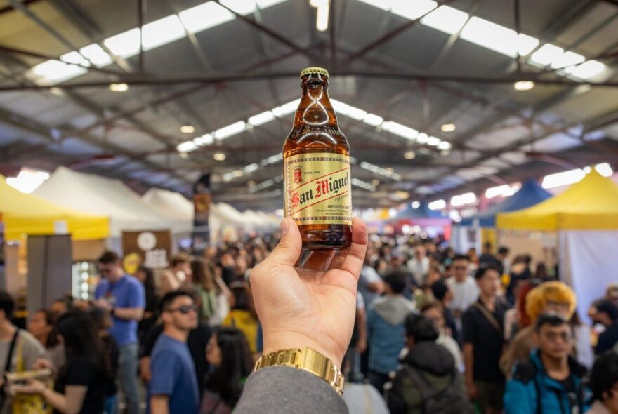 A bottle of San Miguel beer raised at market event Pinoy Octoberfest, held at the Queen Victoria Market, with a blurred crowd in the background.