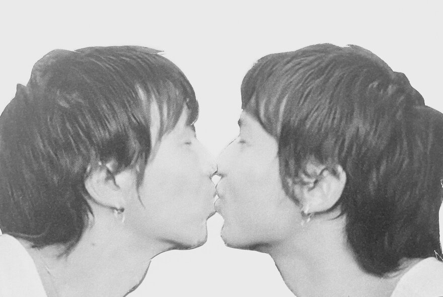 A black and white image of a person kissing a mirror so it looks like they're kissing themselves on the lips. 