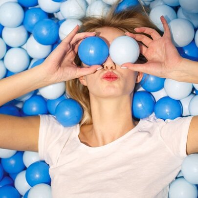 Person lying on a bed of blue plastic balls, holding two balls up to her eyes.