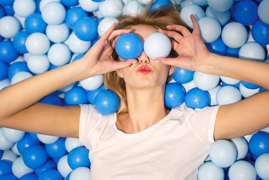 Person lying on a bed of blue plastic balls, holding two balls up to her eyes.