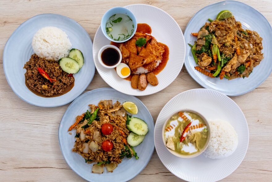 Several dishes of Thai food on a light wooden table, including stir-fried rice and noodles.