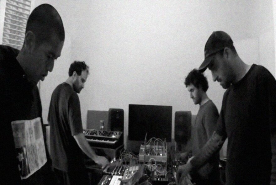 Four people working together on electronic music production, all of them interacting with equipment; black and white image.