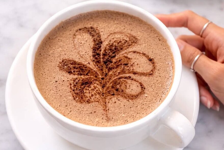 A white cup of coffee or hot chocolate garnished with a cocoa powder stencil in the shape of a flower.