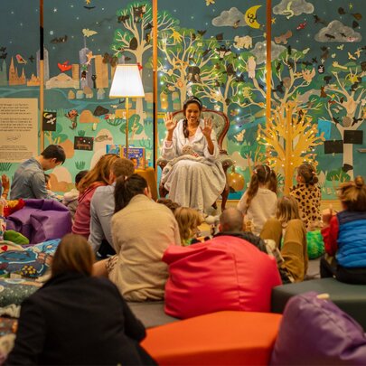 A night-time story session at the State Library Victoria, with young children and their grown-ups listening to a story while sitting on the floor or on beanbags; a colouful mural on the wall behind the story-teller.