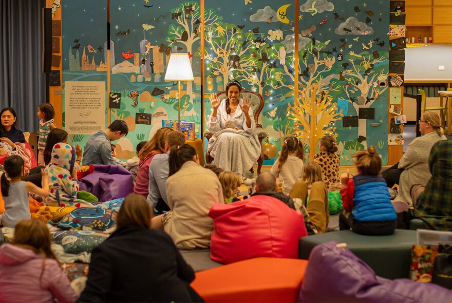 A night-time story session at the State Library Victoria, with young children and their grown-ups listening to a story while sitting on the floor or on beanbags; a colouful mural on the wall behind the story-teller.