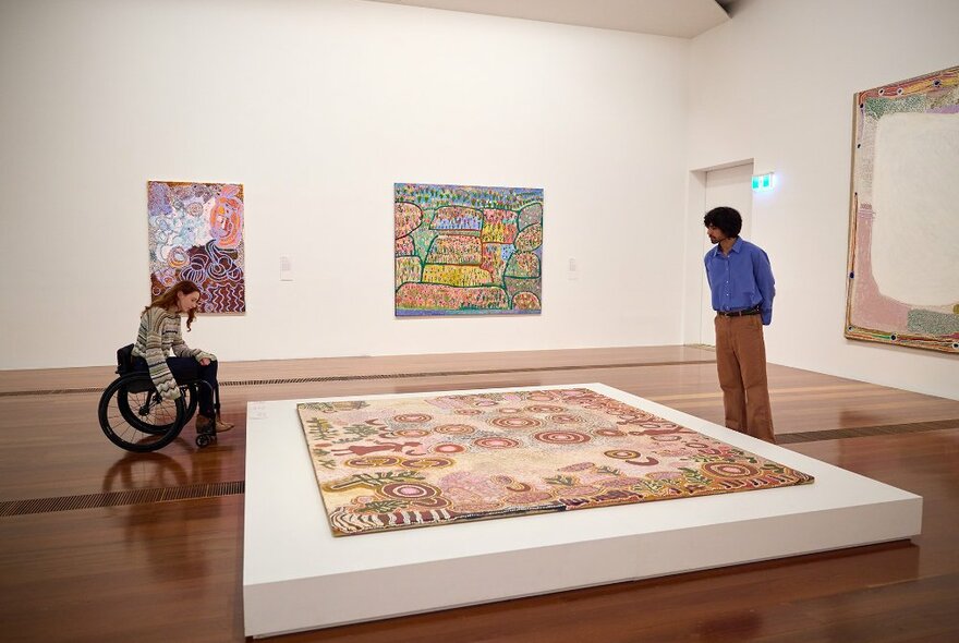 Two people looking at an Aboriginal painting displayed on the floor of a gallery. The woman is using a wheelchair.