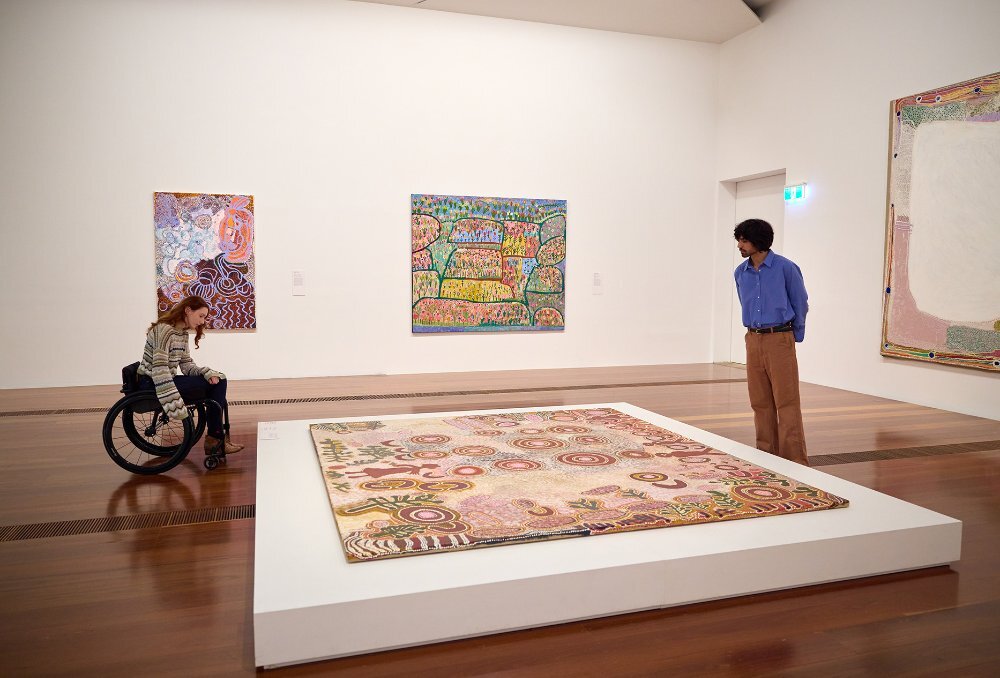 Two people looking at an Aboriginal painting displayed on the floor of a gallery. The woman is using a wheelchair.