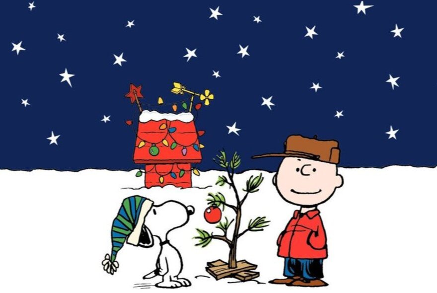 Cartoon of Charlie Brown and Snoopy standing outside in the snow in front of a small Christmas tree, with Snoopy's kennel in the background covered in festive decorations.