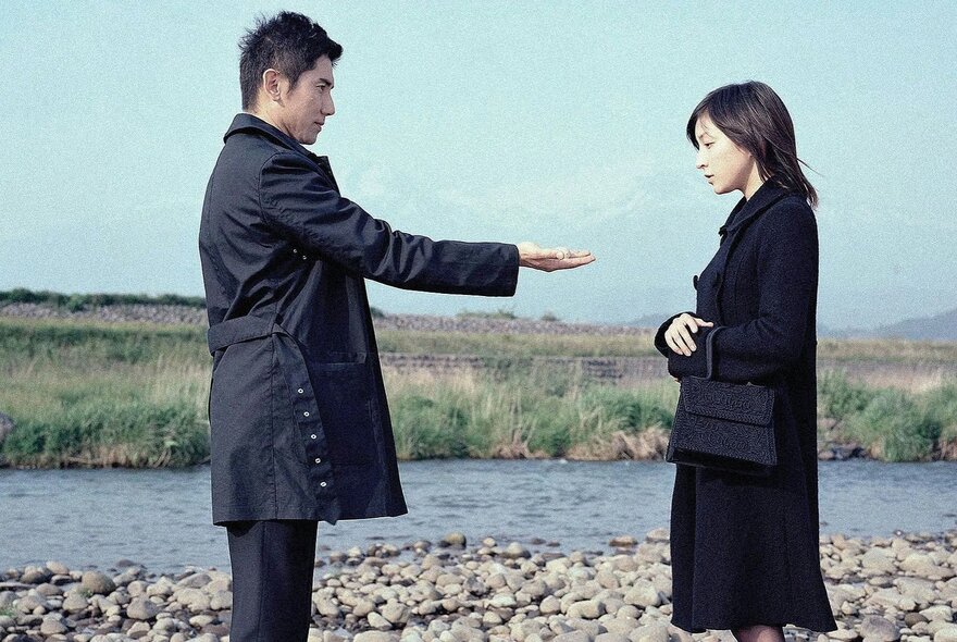 Film still of a man holding out his hand to a woman in a stony river landscape.