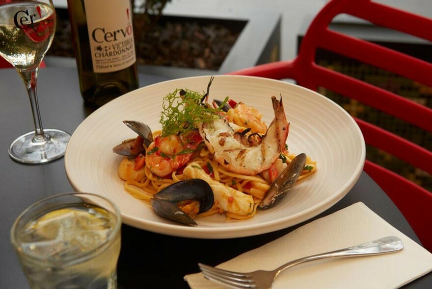 Dish of pasta marinara with prawns, with glass and bottle of white wine, cutlery and water.